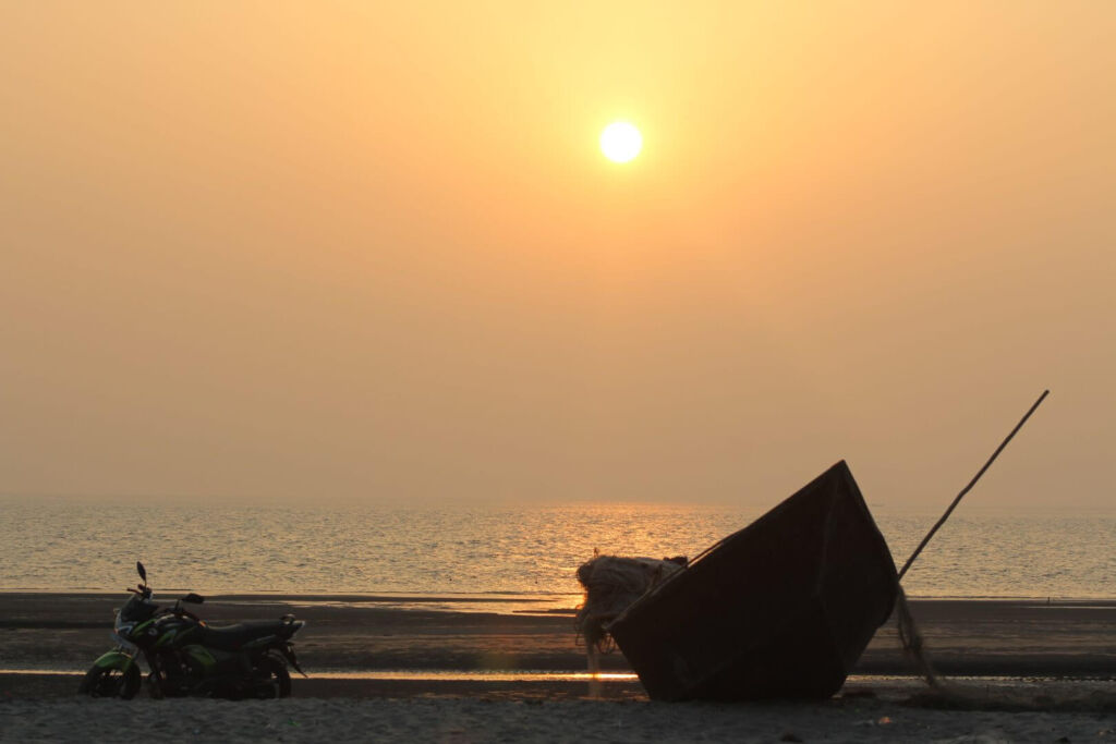 A tranquil sunset bathes the beach, illuminating a parked motorcycle and a beached boat with its fishing net.