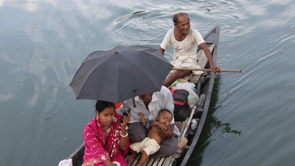 A family travels by boat with an older man propelling them forward, while a woman in a pink sari and a man holding a baby are sheltered under an umbrella.