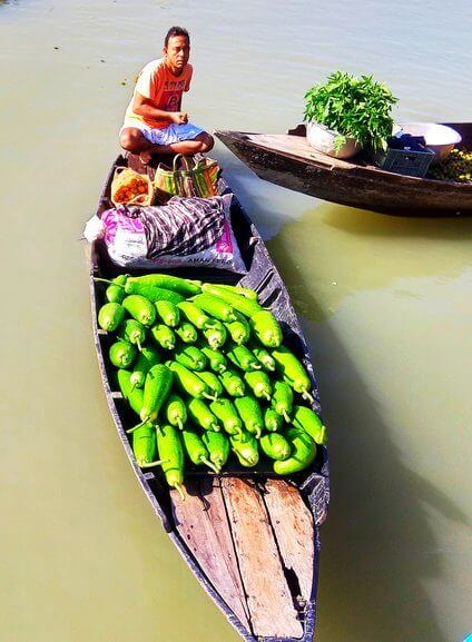 A man crouches on a boat filled with green gourds, likely selling them on the water alongside another boat with plants.