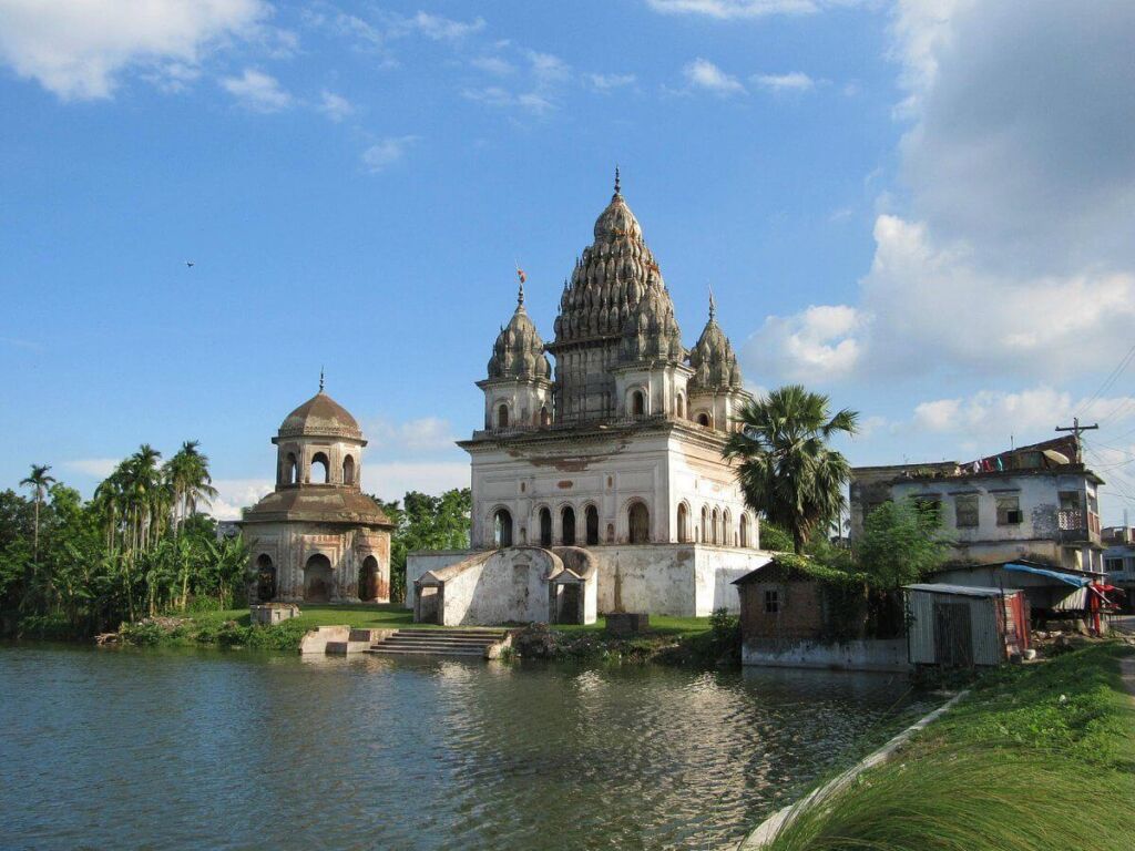 This image showcases a picturesque scene of an old temple complex with ornate architecture, reflected in the calm water of a pond, under a bright blue sky with scattered clouds.