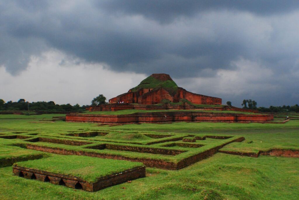 An ancient brick structure, likely a Buddhist monastery, sits amidst lush green grass under a cloudy sky, hinting at a rich historical and cultural landscape.