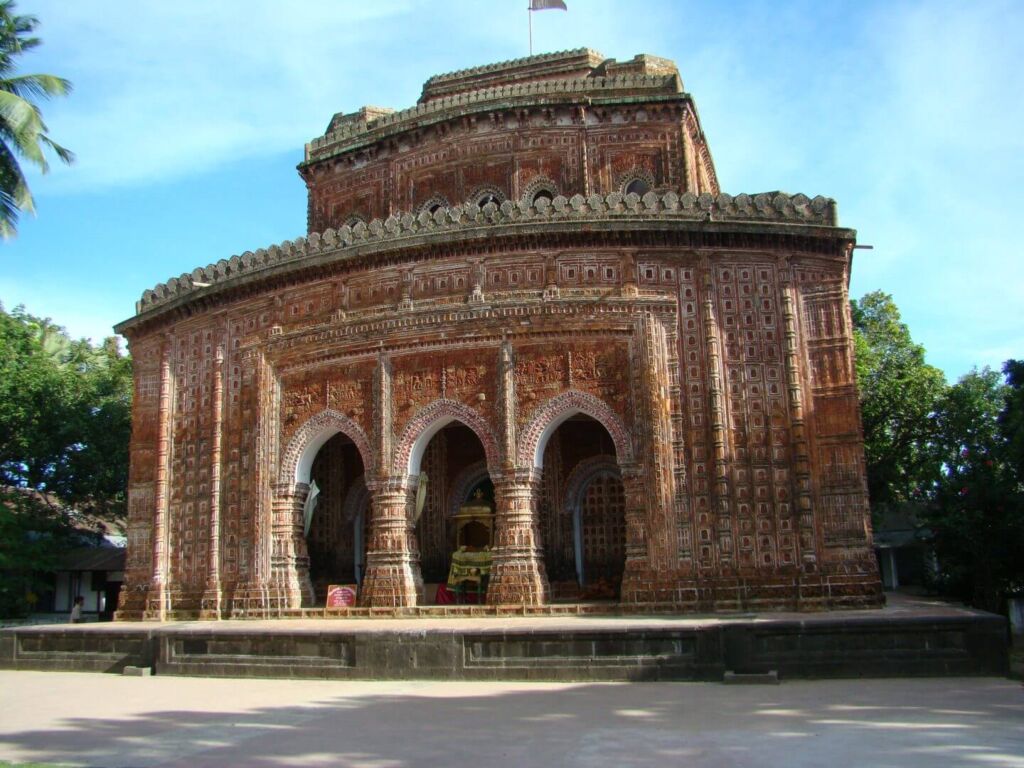 A red brick temple with intricate carvings stands on a raised platform under a blue sky, framed by trees.
