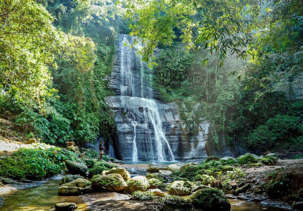 A scenic view of a tiered waterfall cascading down a rock face, surrounded by lush green vegetation and mossy rocks, with a person standing near the base.