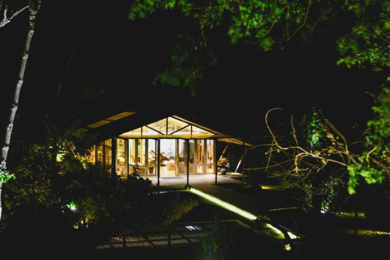 An illuminated glass house stands amidst darkness, surrounded by trees and greenery, with a light path leading to its inviting entrance.