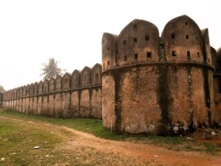 Sonargaon Old Capital Tour An old, weathered stone fortress wall with crenellations and a round tower stands along a dirt path, under an overcast sky with a distant palm tree.