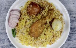 Dhaka Food Workshop A plate of chicken biryani with a chicken drumstick, a piece of meat, a boiled egg, sliced onions, and green chilies.