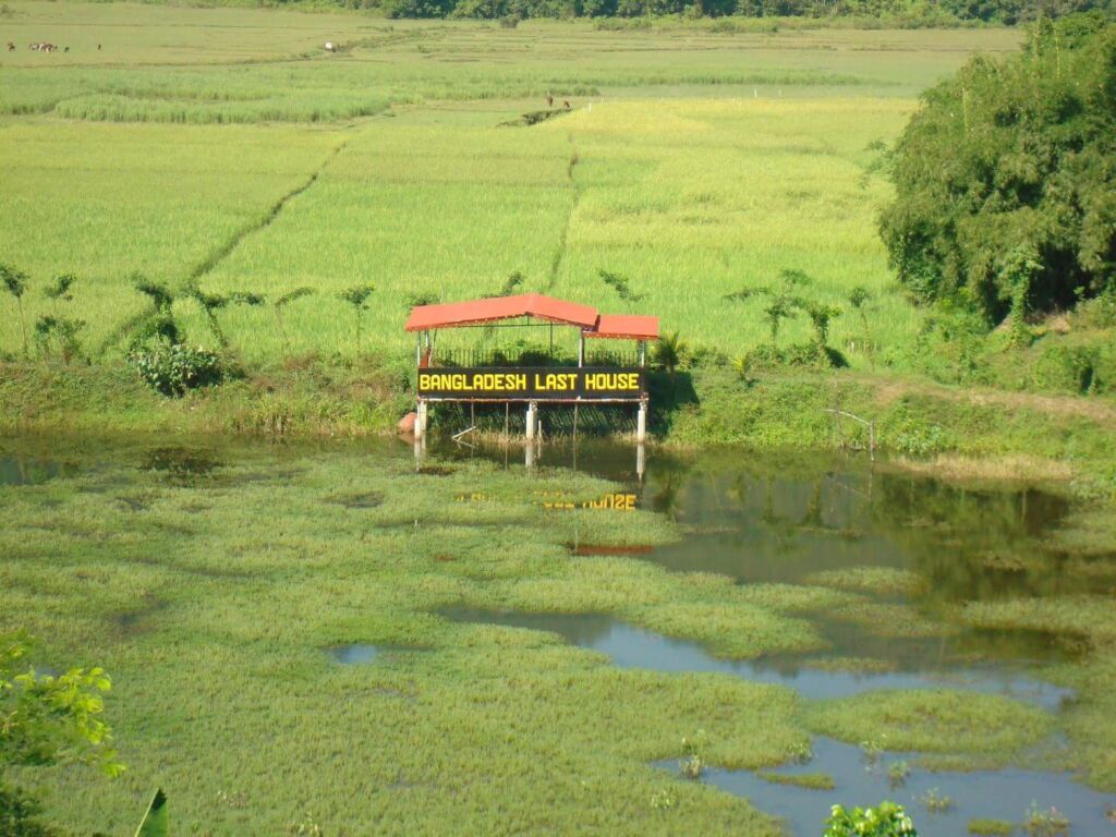 A landscape featuring a green rice field, a body of water covered in vegetation, and a small structure with a red roof labeled 