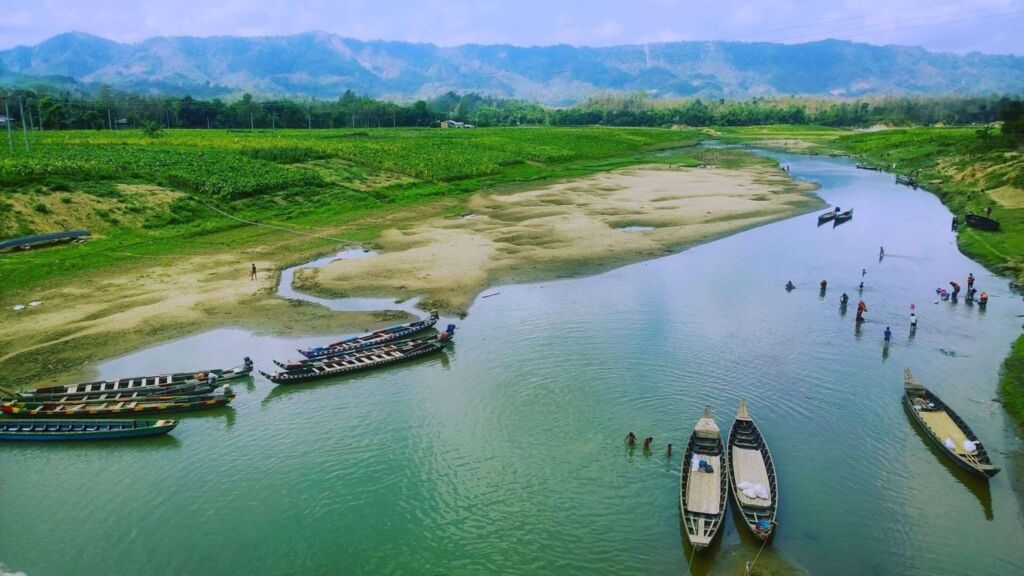 A serene river scene with boats, people wading in the water, and lush green landscapes and mountains in the background.