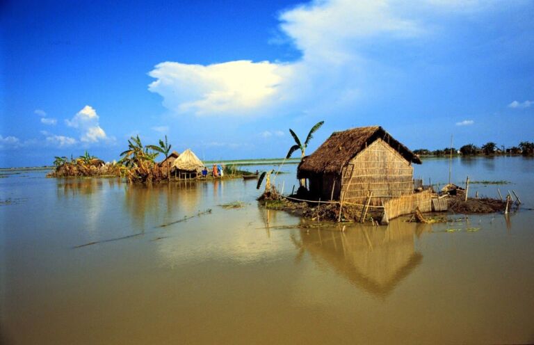 A flooded landscape shows a thatched-roof house and other structures partially submerged in water under a blue, cloudy sky, likely indicating a severe flood event.