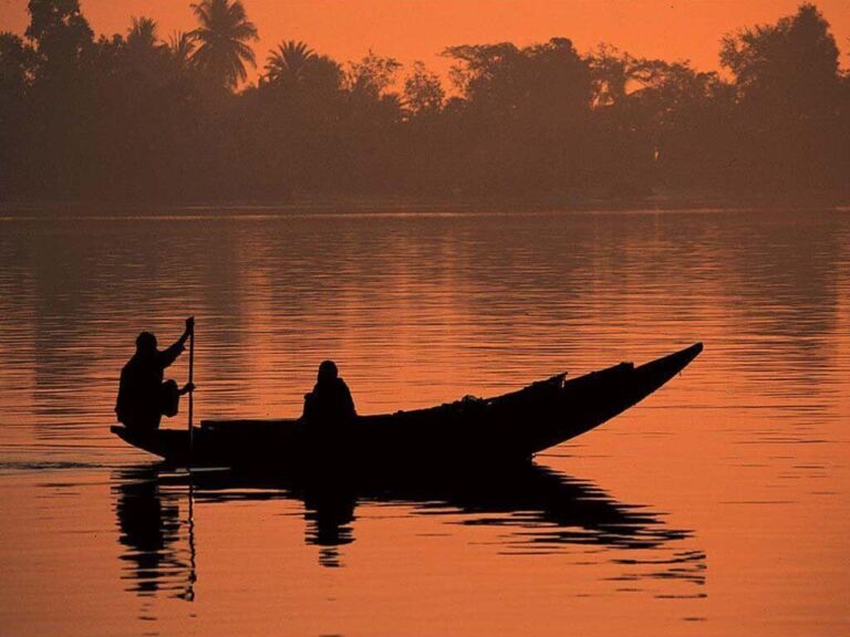 A silhouette of two people in a boat on a calm body of water during an orange sunset.