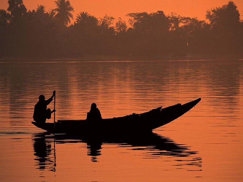 A silhouette of two people in a boat on a calm body of water during an orange sunset.