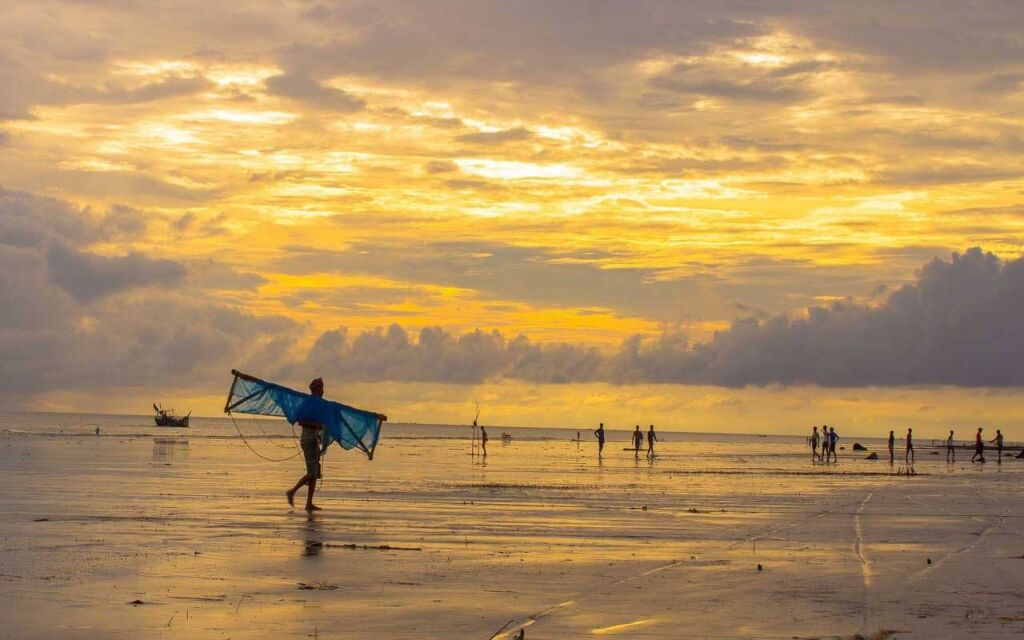 A person carries a net on a beach with a golden sunset, reflecting on the wet sand, with silhouettes of people and a boat in the distance.
