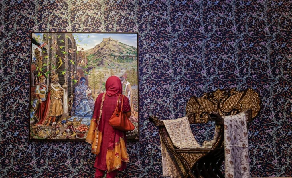A woman in a red hijab and tunic stands in front of a painting in a room with patterned wallpaper, looking at the artwork depicting figures in a landscape.
