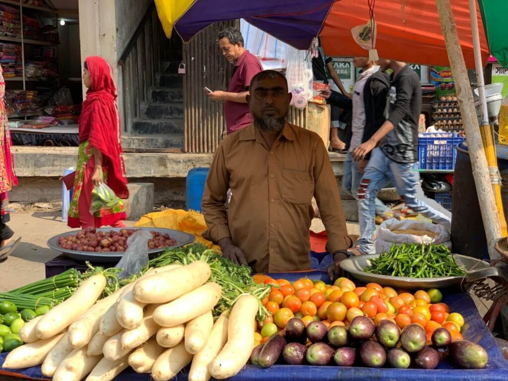 A vendor stands behind a table laden with radishes, tomatoes, eggplants, and chilies at a bustling outdoor market.