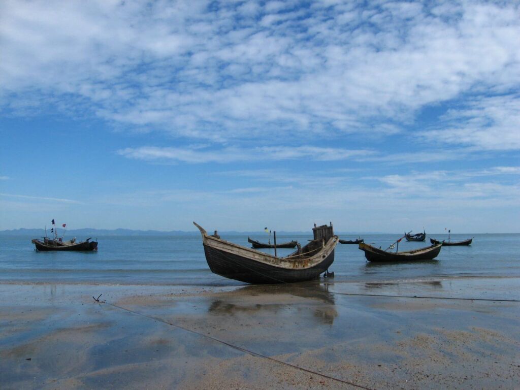 The image shows several wooden boats resting on a wet sandy beach under a bright blue sky dotted with fluffy white clouds.
