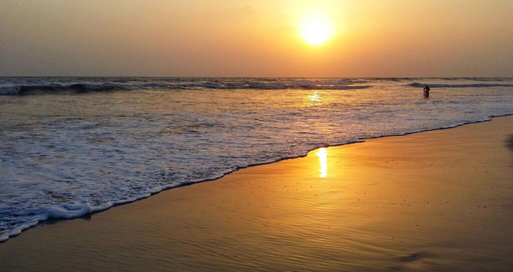 A golden sunset casts a warm glow over the beach and ocean waves, with a silhouette of a person in the distance.