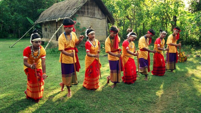 A group of young people in traditional attire dance in a line on a grassy field, holding sticks, with a hut and trees in the background.