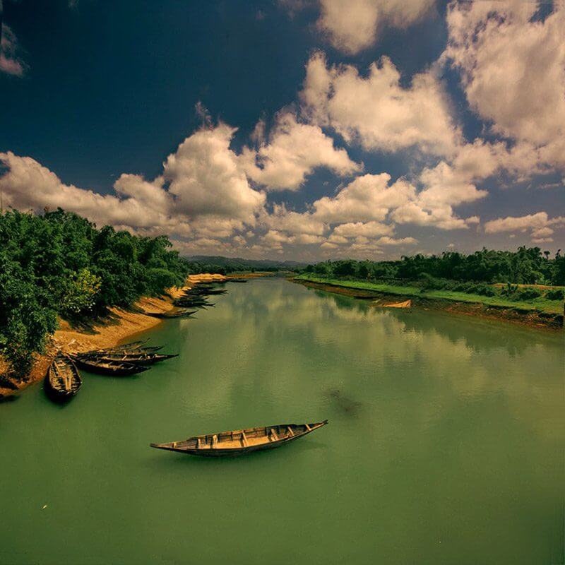 A serene river scene features boats resting on the sandy bank, lush trees lining the shore, and a vibrant sky dotted with fluffy clouds.
