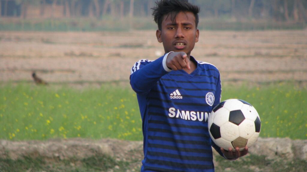 A young man in a blue Chelsea soccer jersey points at the viewer while holding a soccer ball in a field.