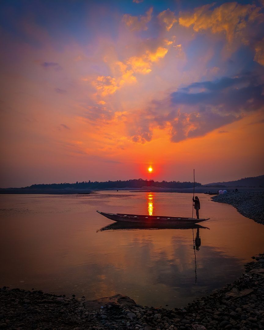 A tranquil sunset over a river with a silhouetted person on a boat, reflecting the vibrant sky.