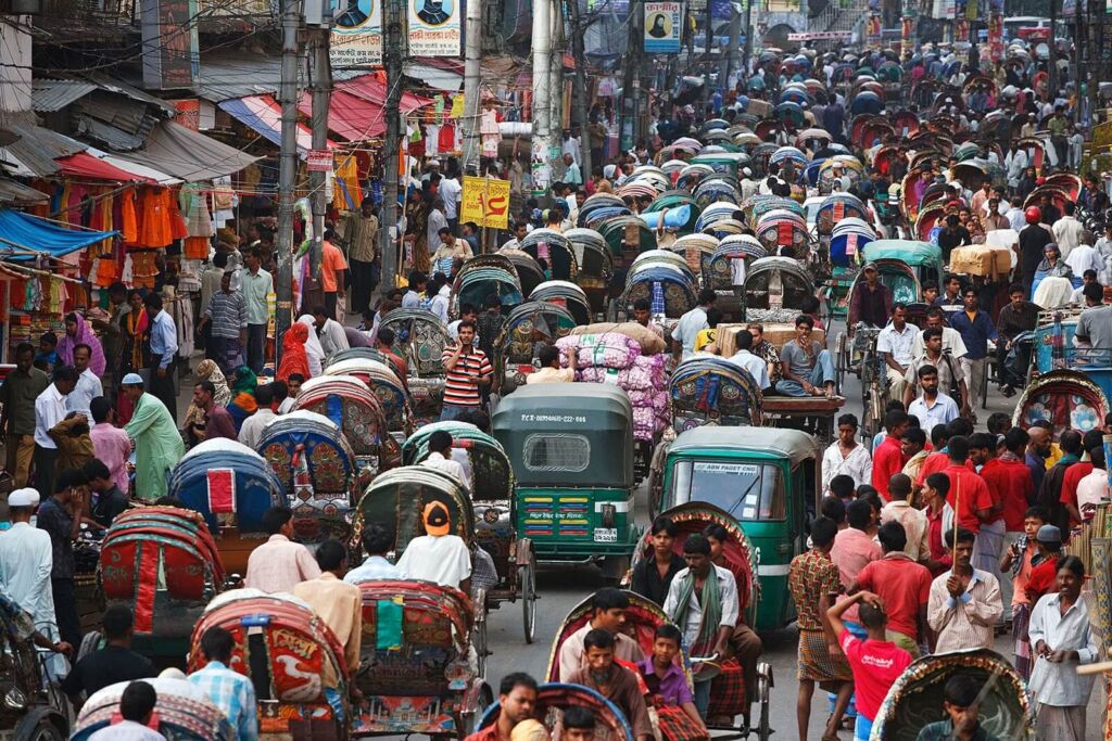 A crowded street scene in Bangladesh features a dense traffic jam of colorful rickshaws and pedestrians, reflecting the bustling urban environment of Dhaka.
