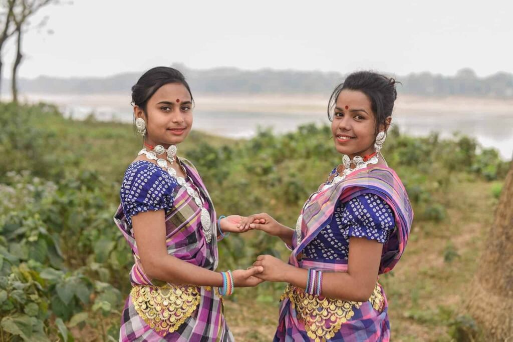 Two smiling women in traditional Indian attire, complete with jewelry and colorful saris, hold hands against a natural backdrop.