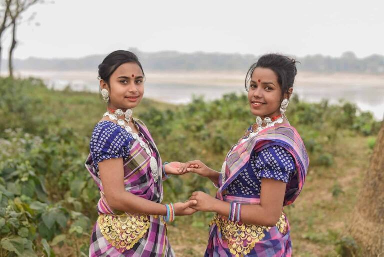 Two smiling women in traditional Indian attire, complete with jewelry and colorful saris, hold hands against a natural backdrop.
