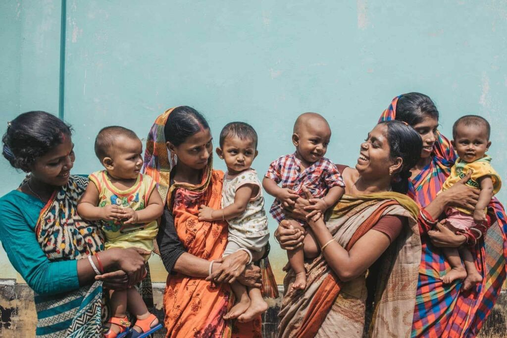 Six Indian women in colorful saris stand in a row, each holding a baby, with a light blue wall behind them.