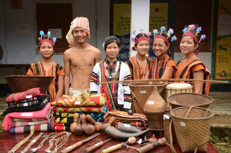 A group of smiling young people, likely from an indigenous community, display their traditional handicrafts such as woven baskets, textiles, and tools on a table.