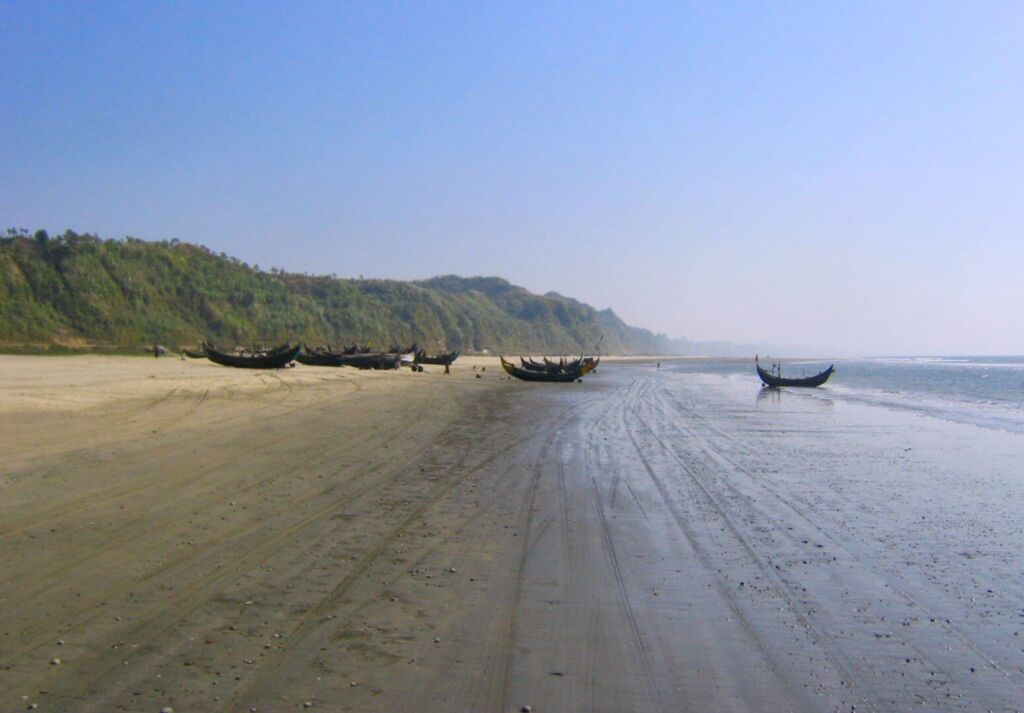 A serene beach scene with multiple traditional boats resting on the sand near lush green hills under a clear blue sky.