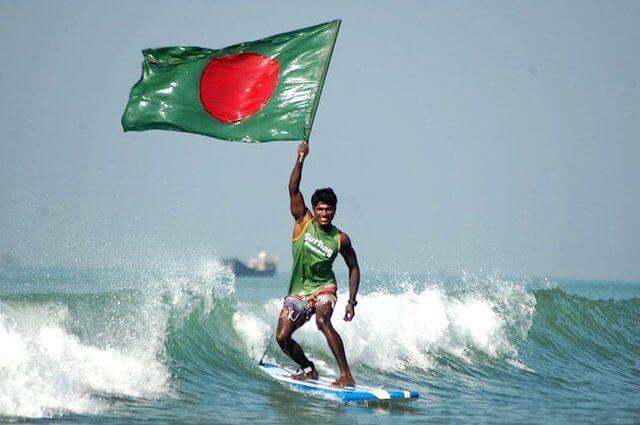 A surfer holds up the flag of Bangladesh while riding a wave.