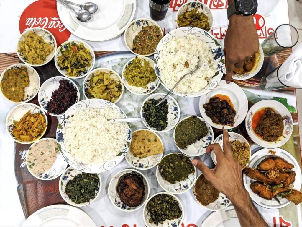 A table laden with many bowls and plates filled with a variety of dishes, including rice, curries, and vegetables, with two hands pointing to different items.