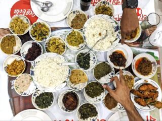 Dhaka Food Tour A table laden with many bowls and plates filled with a variety of dishes, including rice, curries, and vegetables, with two hands pointing to different items.
