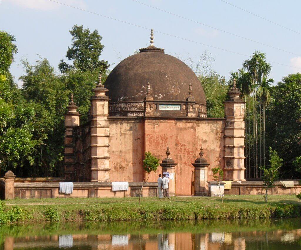 A mosque with a large dome is reflected in a pond, surrounded by trees and greenery.