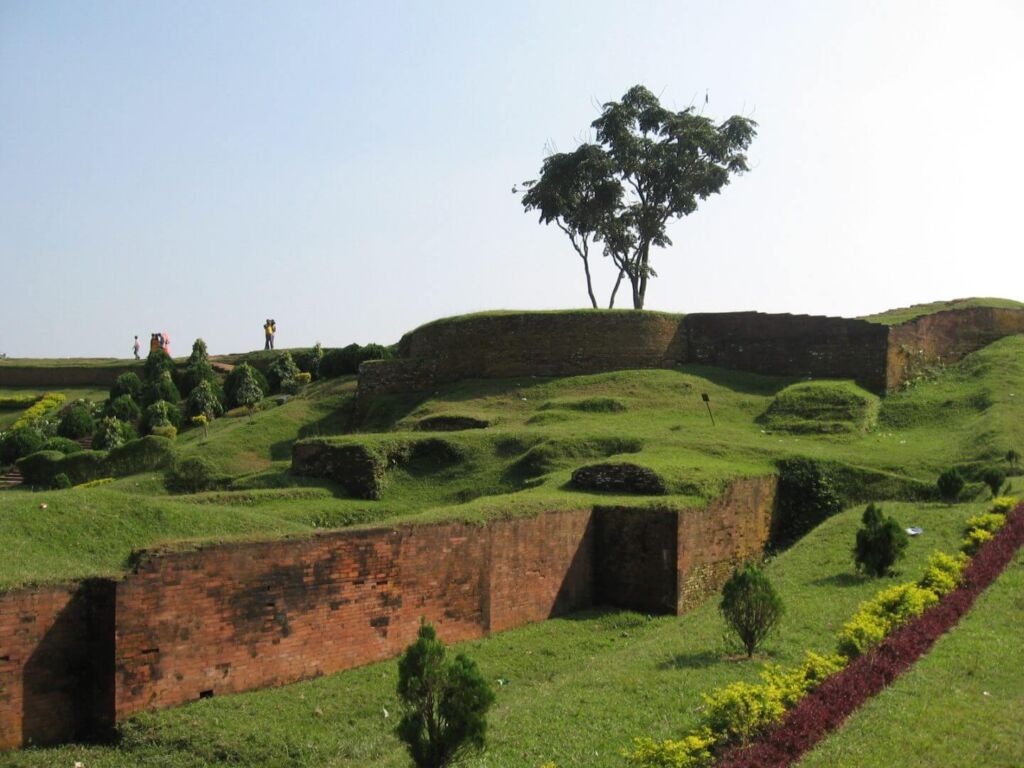 A scenic landscape of a grassy hill with brick walls, manicured bushes, a solitary tree at the top, and people walking in the distance against a clear sky.