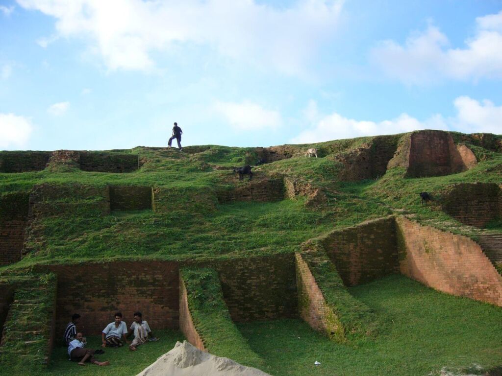 People and goats populate an ancient brick structure covered in grass, set against a blue sky with scattered clouds.