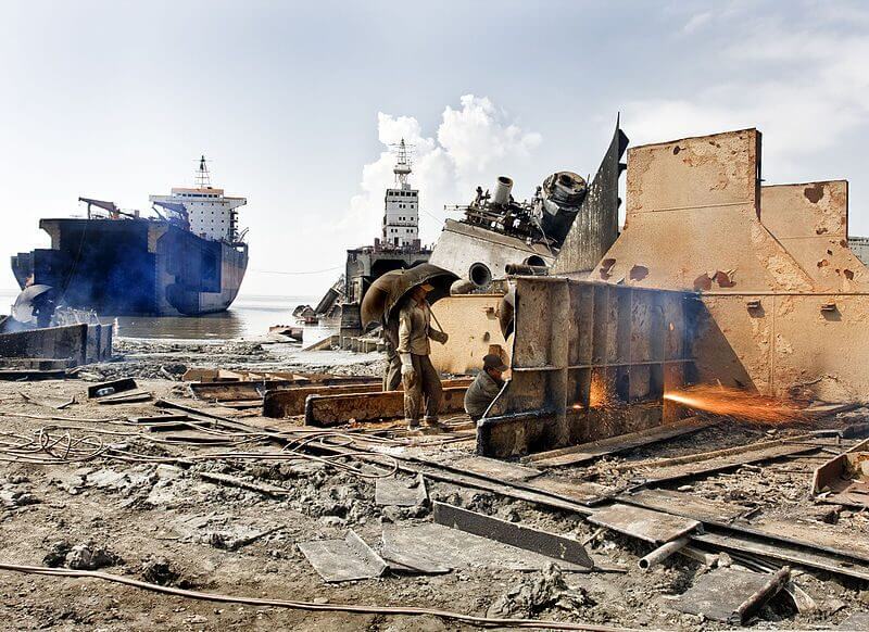 Workers dismantle a ship in a shipyard, with one welding metal and another carrying materials under an umbrella amidst the debris.