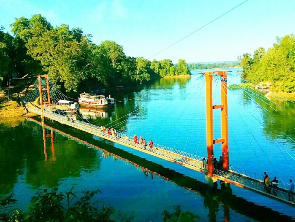 A suspension bridge stretches across a tranquil river, connecting lush green shores, with boats anchored nearby and people strolling across under a bright blue sky.