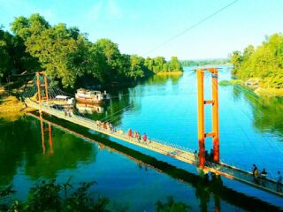 Kaptai Lake Day Tour A suspension bridge stretches across a tranquil river, connecting lush green shores, with boats anchored nearby and people strolling across under a bright blue sky.