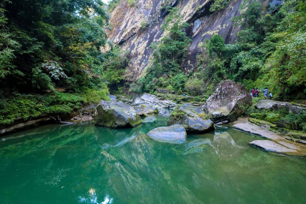 A tranquil emerald pool surrounded by mossy rocks and lush greenery, nestled at the base of a steep cliff face with a group of people on the right.