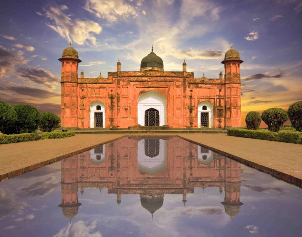A red building with domes is reflected in a pool of water under a cloudy sky.