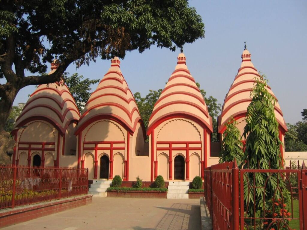 A captivating view of four traditional Bengali Hindu temples with conical roofs, adorned with red stripes, set within a fenced garden.