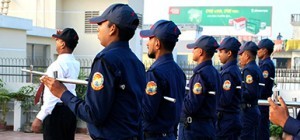 A line of security guards in dark blue uniforms and caps stand at attention, some holding batons, with a man in a white shirt and tie in front.