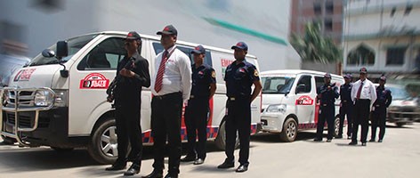 A line of security guards in uniform stands in front of two white vans with red logos.