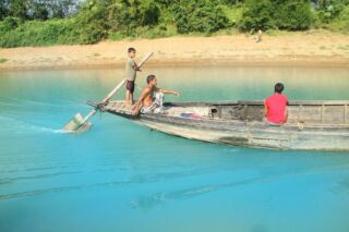 Lalakhal And Jaflong Three people are on a weathered wooden boat in a river with vibrant turquoise water, with two standing near the bow, one seated in the back, and vegetation in the background.