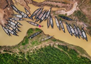 Ratargul And Bholaganj Sada Pathor An aerial view shows numerous traditional wooden boats gathered in a muddy waterway, surrounded by patches of green vegetation and earth.