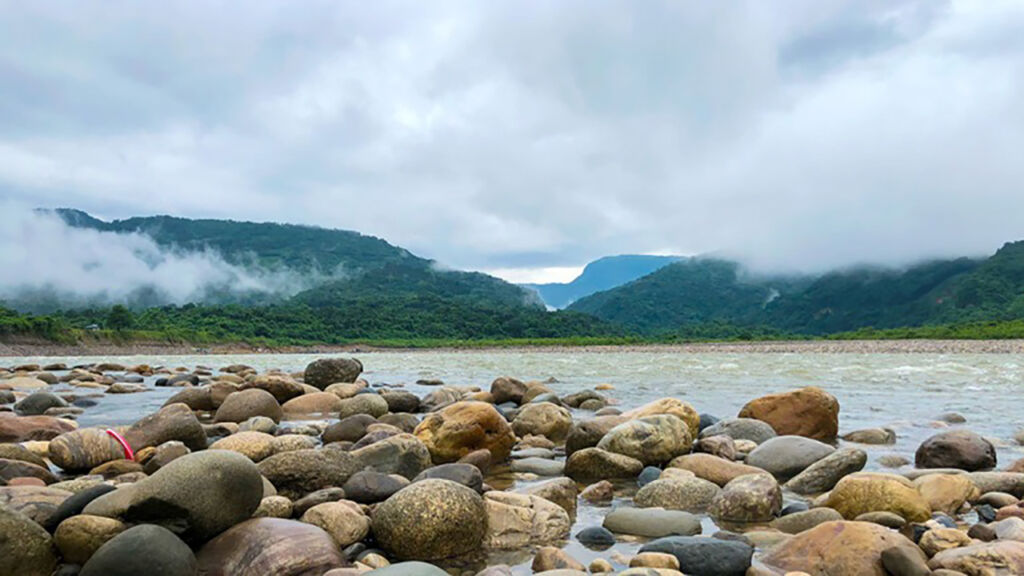 A scenic view of a rocky riverbank leading to flowing water, surrounded by lush green hills partially covered by clouds under an overcast sky.