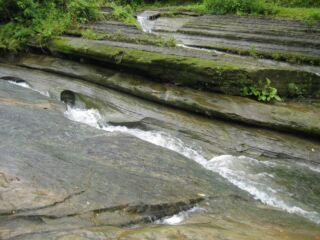 A stream cascades over layered rock formations covered in moss and greenery, creating a natural and scenic landscape.