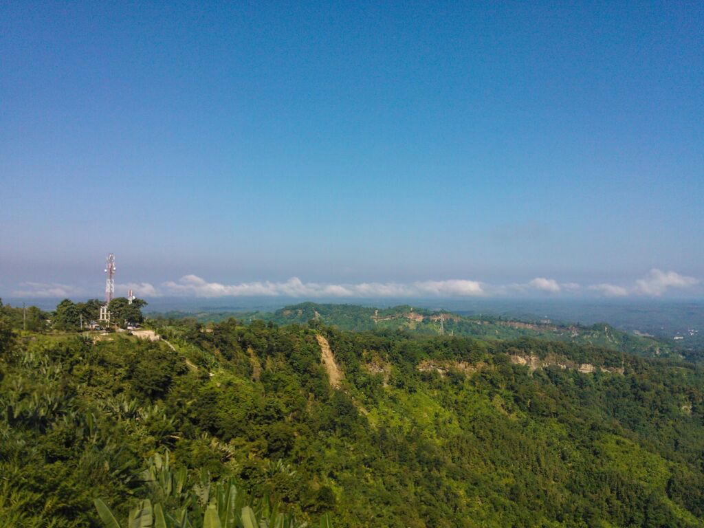 A lush green hillside, dotted with trees, descends from a hilltop with a communications tower, under a clear blue sky with a distant band of white clouds.