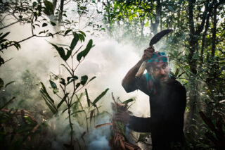 A man in a forest stands amidst smoke, holding a sickle near his head.